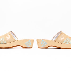 May include: A pair of beige slip-on clogs with a low platform heel and a brown rubber sole. The upper features light blue and peach-colored fish illustrations, while the inner lining is silver. The side of the heel also displays a light blue fish graphic.