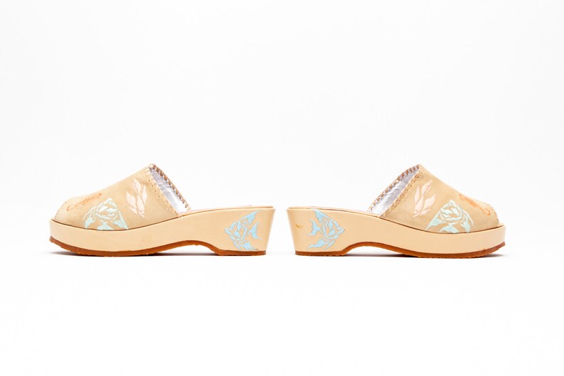May include: A pair of beige slip-on clogs with a low platform heel and a brown rubber sole. The upper features light blue and peach-colored fish illustrations, while the inner lining is silver. The side of the heel also displays a light blue fish graphic.