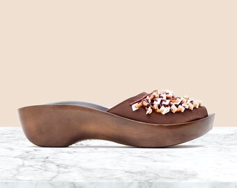 May include: A brown slip-on mule with a thick, contoured wooden platform sole. The upper is made of brown fabric adorned with a cluster of small, multi-colored beads and white square-shaped embellishments across the top of the foot area.