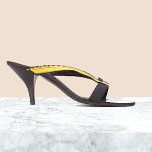 May include: Black heeled sandals with a yellow zipper detail. The sandals have a thin strap across the toes and a thicker strap around the ankle.