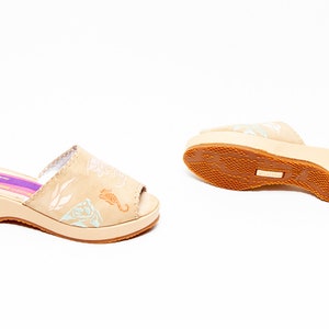 May include: A pair of beige open-toe slide sandals with a low wedge heel. The upper features light blue and white marine-themed illustrations including a seahorse. A purple label inside the footbed displays the text MADE IN ITALY.