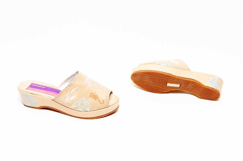 May include: A pair of beige open-toe slide sandals with a low wedge heel. The upper features light blue and white marine-themed illustrations including a seahorse. A purple label inside the footbed displays the text MADE IN ITALY.