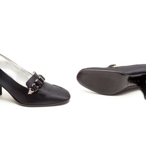 May include: A pair of black satin pumps featuring a block heel and a decorative strap across the vamp adorned with rectangular black crystals. The interior lining is silver, and the soles are black with a visible brand stamp on the bottom.