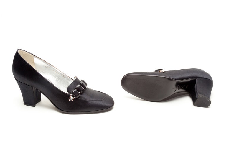 May include: A pair of black satin pumps featuring a block heel and a decorative strap across the vamp adorned with rectangular black crystals. The interior lining is silver, and the soles are black with a visible brand stamp on the bottom.
