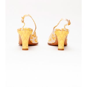 Puede incluir: Un par de tacones amarillos y blancos con tira en el tobillo y un estampado floral. Los tacones miden aproximadamente 7,5 cm de alto.