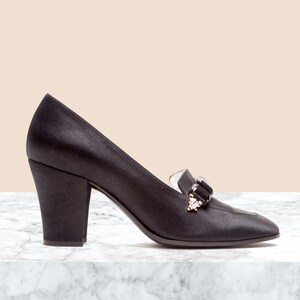 May include: A black satin pump with a block heel and a pointed toe. The shoe features a decorative embellishment on the vamp consisting of a large black rectangular stone flanked by smaller clear crystals set in a gold-tone metal base.