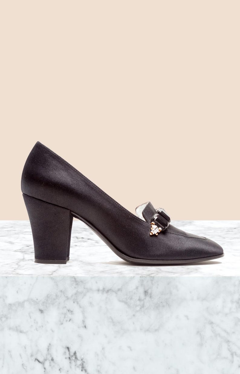 May include: A black satin pump with a block heel and a pointed toe. The shoe features a decorative embellishment on the vamp consisting of a large black rectangular stone flanked by smaller clear crystals set in a gold-tone metal base.