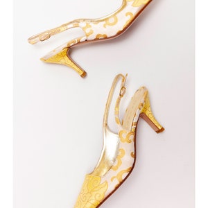 Puede incluir: Un par de tacones amarillos y blancos con tira en el tobillo y estampado floral. Los tacones son de punta y tienen una tira delicada alrededor del tobillo.