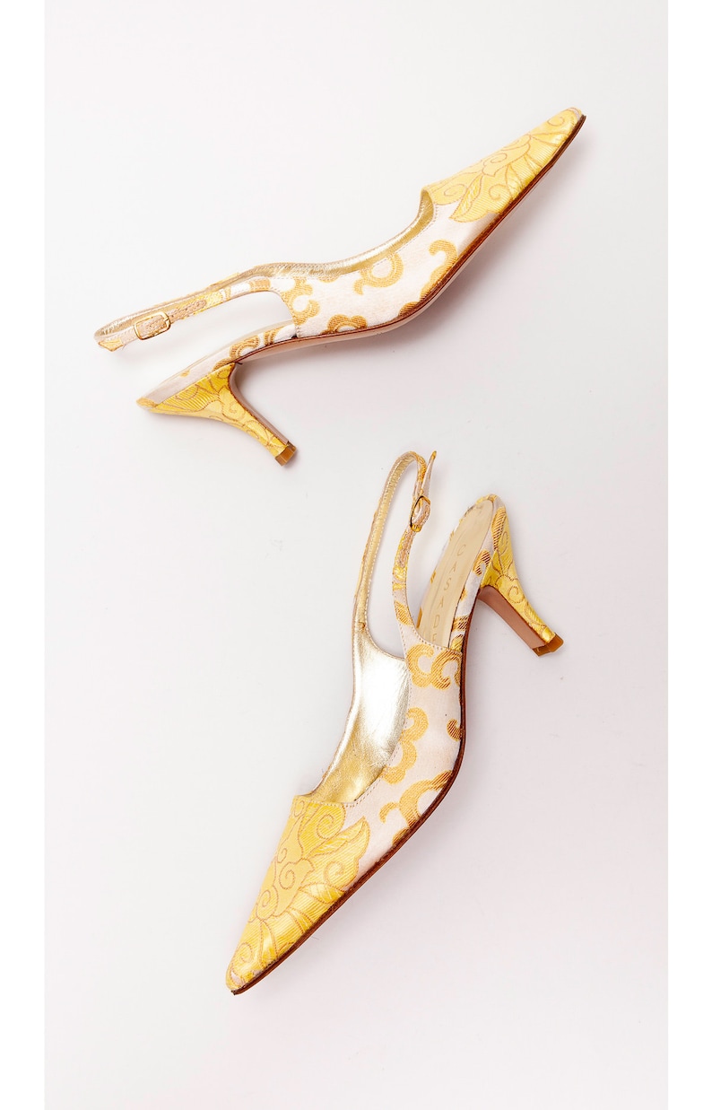 Puede incluir: Un par de tacones amarillos y blancos con tira en el tobillo y estampado floral. Los tacones son de punta y tienen una tira delicada alrededor del tobillo.