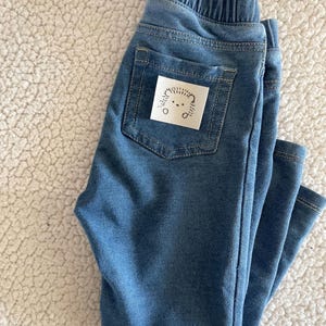 May include: Blue denim trousers with a small white patch on the back pocket featuring a black and white illustration of a hedgehog.