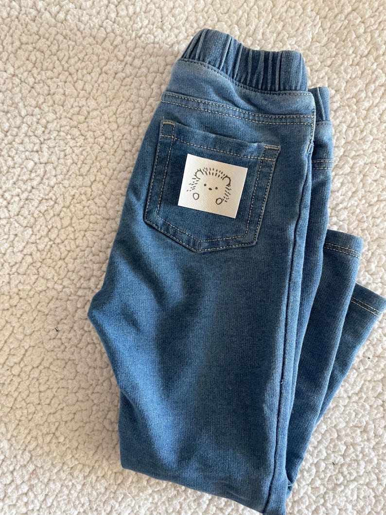 May include: Blue denim trousers with a small white patch on the back pocket featuring a black and white illustration of a hedgehog.