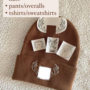 May include: Brown knitted beanie with white embroidered antlers and a blank space for a transfer. The image also shows four iron-on transfers with animal designs: a deer, a fox, a hedgehog, and another set of antlers.