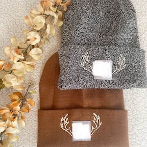May include: Two beanies, one gray and one brown, are displayed. Both beanies have a white embroidered antler design with a square patch. The beanies are folded at the brim and rest on a white, textured surface.