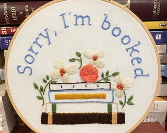 May include: Embroidered hoop art featuring the text "Sorry, I'm booked" in blue thread. The design includes a stack of books with a floral arrangement on top. The books are brown, green, and blue with a yellow spine. The flowers are white and pink.