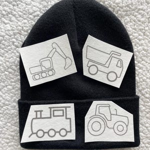 May include: Black beanie with four white patches featuring outlines of a tractor, a dumper truck, an excavator, and a train. The patches are blank and ready to be coloured in.