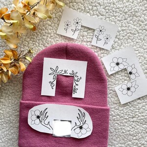May include: A pink beanie with a white patch featuring a floral design and antlers. The patch has a rectangular cutout in the centre. There are four additional white patches with floral designs.