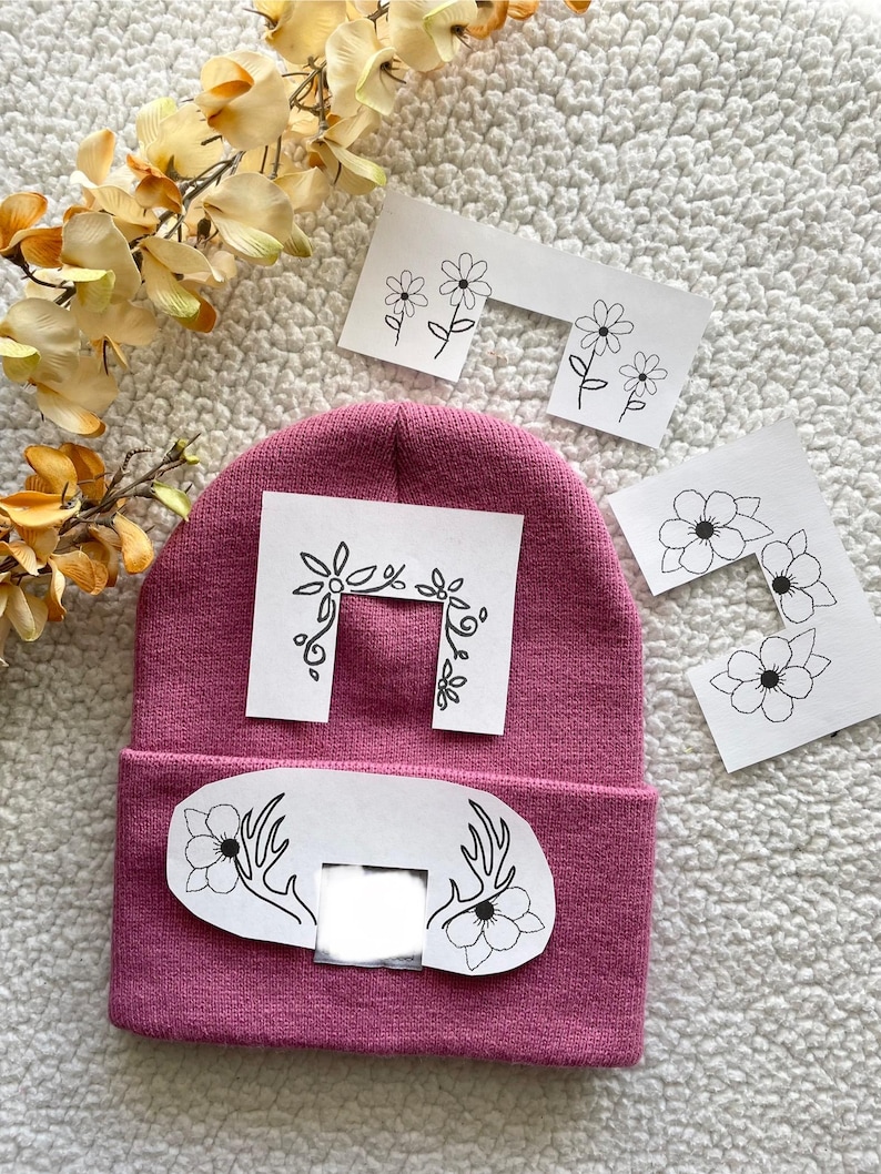 May include: A pink beanie with a white patch featuring a floral design and antlers. The patch has a rectangular cutout in the centre. There are four additional white patches with floral designs.