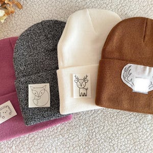 May include: Four knitted beanies in different colours with embroidered animal designs. The beanies are pink, grey, white, and brown. The designs include a fox, a deer, and a set of antlers.