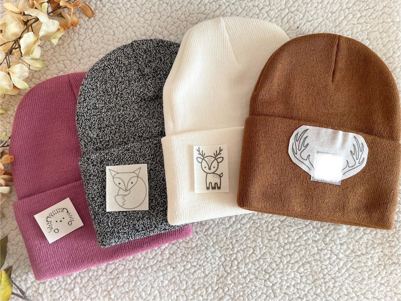May include: Four knitted beanies in different colours with embroidered animal designs. The beanies are pink, grey, white, and brown. The designs include a fox, a deer, and a set of antlers.