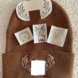 May include: Brown knitted beanie with a white deer applique and a white deer antler applique. There are three additional white appliques with a deer, a fox, and a hedgehog.