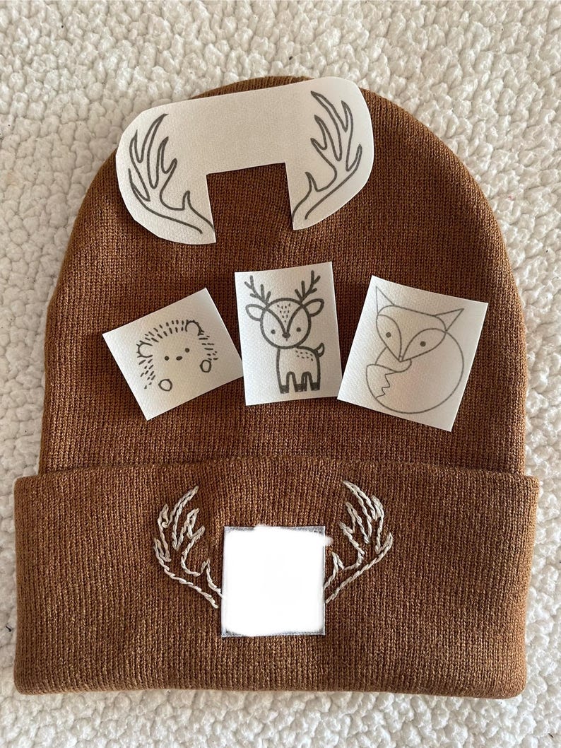 May include: Brown knitted beanie with a white deer applique and a white deer antler applique. There are three additional white appliques with a deer, a fox, and a hedgehog.
