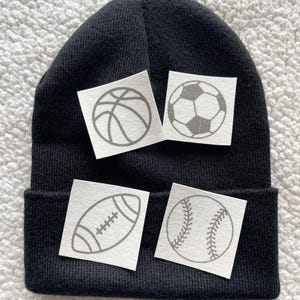 May include: Black beanie with four white patches featuring different sports ball designs: basketball, football, rugby ball, and baseball.