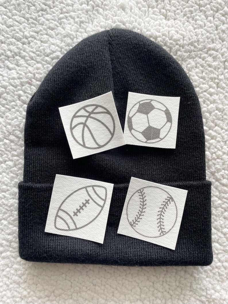May include: Black beanie with four white patches featuring different sports ball designs: basketball, football, rugby ball, and baseball.