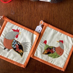 Könnte beinhalten: Zwei quadratische, cremefarbene Topflappen mit orangefarbenem Rand. Jeder hat eine farbenfrohe Hahnapplikation. Ein Hahn blickt nach links, der andere nach rechts. Die Hahn-Designs umfassen rote, graue, orangefarbene, grüne und schwarze Stoffdetails.