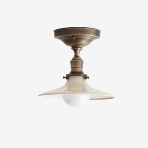 May include: A vintage-style ceiling light fixture with a brass finish. The light has a white, saucer-shaped shade and a round, white bulb. The fixture is designed for direct ceiling mounting and adds a touch of classic elegance.