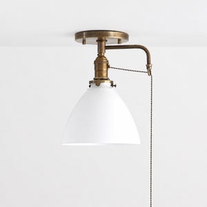 May include: A ceiling-mounted light fixture featuring a matte white conical shade and an antique brass finish metal arm. The fixture includes a vertical pull chain mechanism attached to the side, allowing for manual operation of the light.