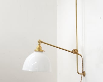 May include: A brass wall-mounted lamp with a white glass shade. The lamp has a long arm that extends out from the wall and can be adjusted to point in different directions.
