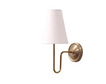 May include: A wall-mounted lamp with a white lampshade and a brass-colored metal frame. The lamp has a curved arm and a circular base. The lampshade is a classic tapered shape. The fixture is designed for indoor lighting.