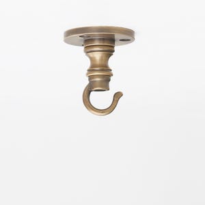 May include: A brass-colored ceiling hook with a circular mounting plate and a curved hook. The hook is designed for hanging items and has a decorative, vintage aesthetic. The mounting plate has three screw holes.