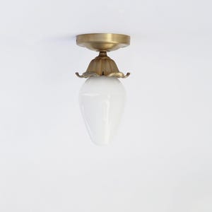 May include: A vintage-style ceiling light fixture with a white, teardrop-shaped glass shade. The fixture has a brass-colored metal base and decorative floral accents. The light is designed to be mounted on a ceiling.