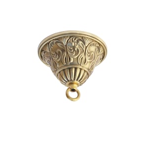 May include: A brass-colored ceiling canopy with ornate floral detailing. The canopy has a bell shape with a ring at the bottom for hanging. The design features a series of vertical lines and decorative patterns.