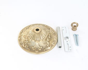 May include: A brass-colored ceiling fixture canopy with ornate floral detailing. Includes a silver rod, a white mounting bracket, a brass-colored loop, and two silver screws. The canopy is approximately 5 inches in diameter.