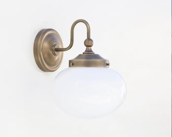 Op de afbeelding: Een wandlamp in vintage stijl met een messing afwerking. De lamp heeft een ronde achterplaat, een gebogen arm en een bolvormige witte glazen kap. Deze lamp is geschikt voor binnen of buiten en voegt een klassieke uitstraling toe.
