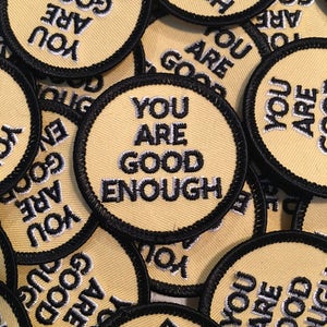 May include: A close-up of several yellow fabric patches with black stitching. The patches are round and have the text "You are good enough" embroidered in white on them.