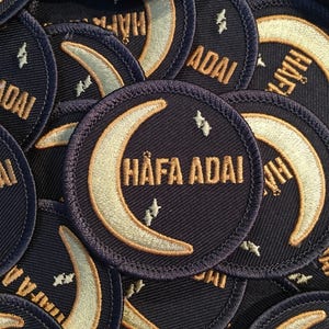 May include: Close-up of several navy blue fabric patches with a crescent moon and stars design. The patches have the text "HAFA ADAI" in gold embroidery.