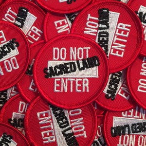 May include: A close-up of several red fabric patches with white and black embroidery. The patches read "Do Not Enter Sacred Land".