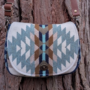 Puede incluir: Un bolso bandolera color crema con un estampado geométrico en tonos azules, marrones y blancos. El bolso tiene un cierre de solapa y correas de cuero marrón. El bolso está colgado de un tronco de árbol.