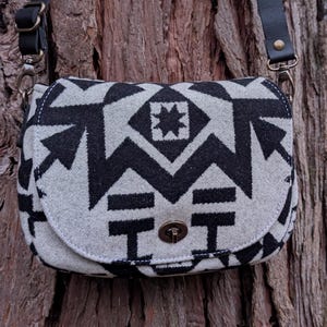 Puede incluir: Bolso bandolera con estampado en blanco y negro y correa de cuero colgado de un árbol. El bolso presenta un diseño geométrico con cierre de solapa y cierre metálico. El patrón del bolso es un diseño repetitivo.