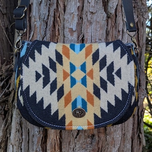Puede incluir: Bolso de montura con un patrón geométrico en crema, azul marino, naranja y turquesa. El bolso tiene una correa de cuero negro y un cierre de metal. El bolso está colgado de un tronco de árbol.