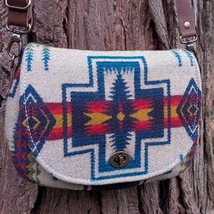 Puede incluir: Bandolera color crema con un estampado geométrico en azul, rojo, amarillo y verde. La bolsa tiene una correa de cuero marrón y un cierre de metal. La bolsa está colgada de un tronco de árbol.