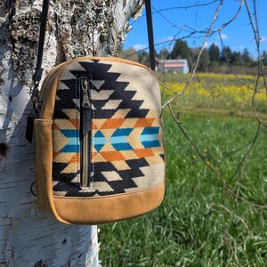 Peut inclure: Un petit sac bandoulière carré avec un motif du sud-ouest en noir, beige, orange et turquoise. Le sac a un fond et des côtés en similicuir beige, une bandoulière noire et une fermeture éclair. Le sac est suspendu à un arbre.