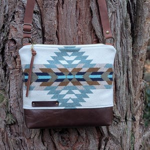 May include: A rectangular crossbody bag with a geometric pattern in shades of blue, brown, and white. The bottom of the bag is brown leather, and it has a brown leather strap. The bag is hanging on a tree.