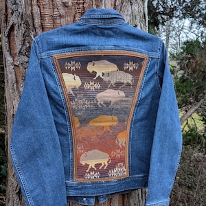 May include: Blue denim jacket with a decorative back panel featuring a bison design in shades of brown, tan, and orange. The jacket is hanging on a metal hanger against a tree trunk background.