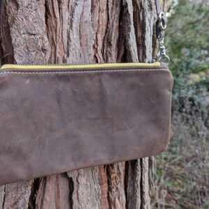May include: A brown leather crossbody bag with a yellow zipper and silver hardware. The bag is rectangular and has a distressed appearance. The strap is brown leather. The bag is hanging on a tree.