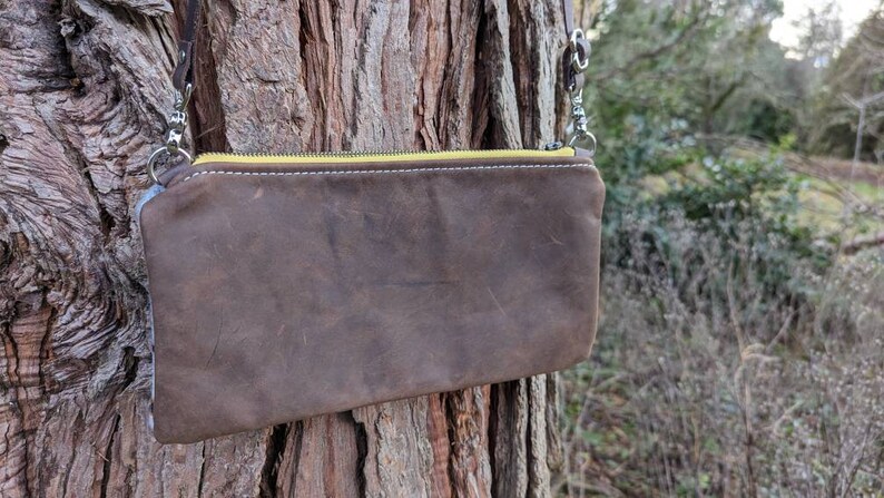 May include: A brown leather crossbody bag with a yellow zipper and silver hardware. The bag is rectangular and has a distressed appearance. The strap is brown leather. The bag is hanging on a tree.