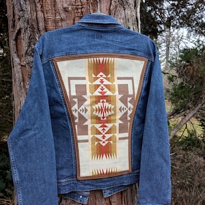 May include: A denim jacket with a decorative back panel. The panel features a geometric pattern in shades of beige, brown, red, and gold. The jacket is hanging on a metal hanger from a tree.